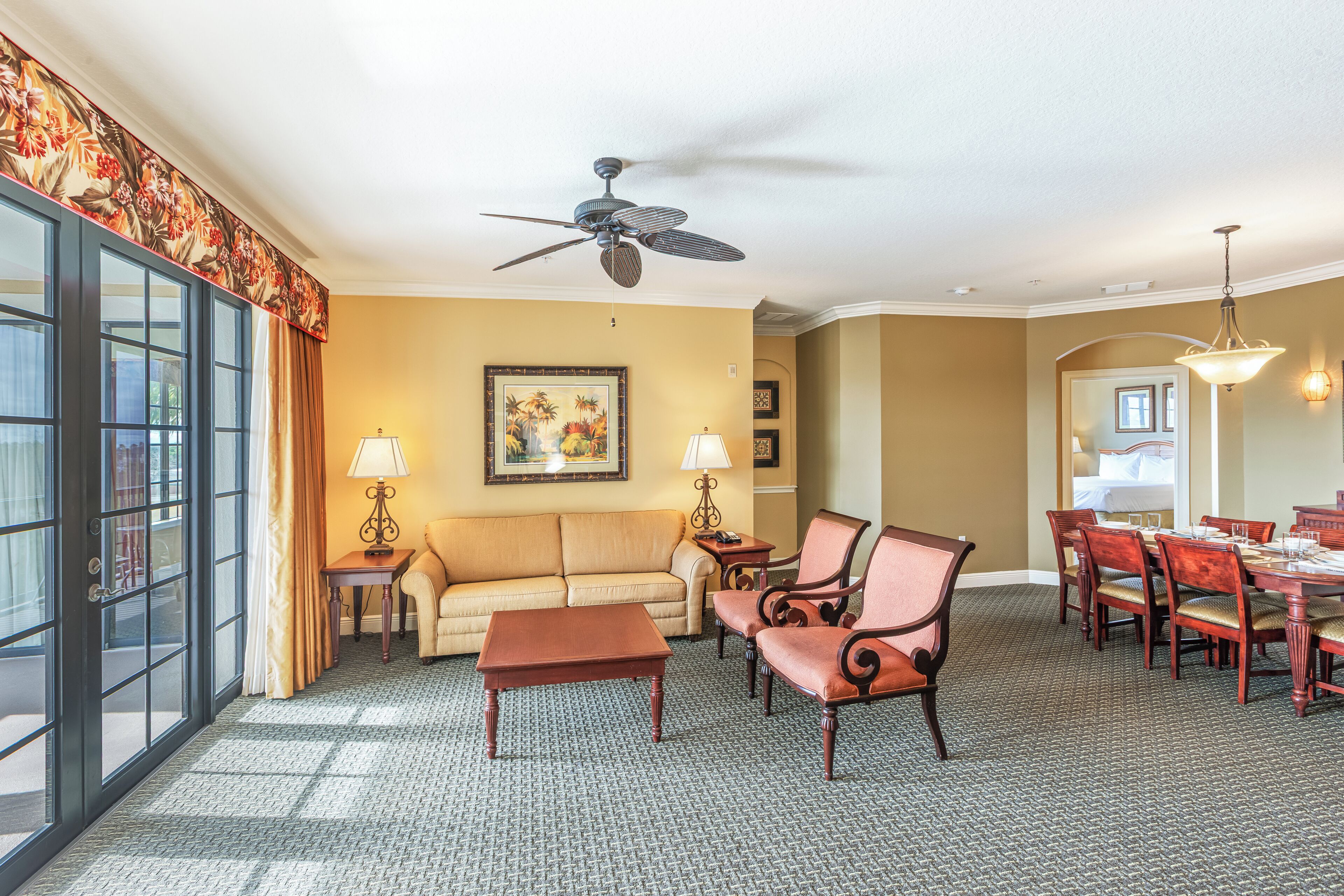 A hotel room with a large window, a ceiling fan, a sofa, a coffee table, a dining table with chairs, and a carpeted floor.