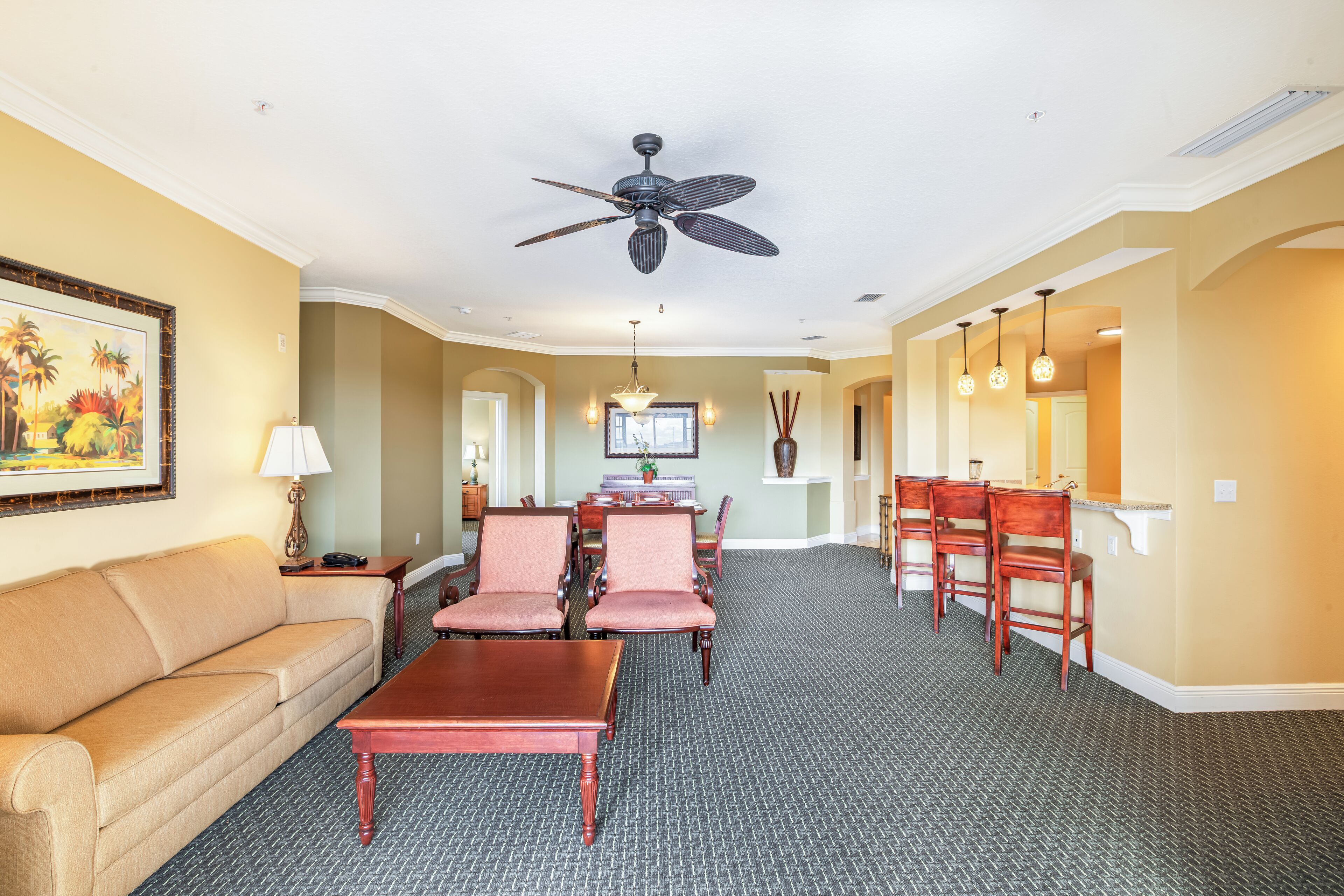 A hotel room with a sofa, dining table, chairs, a ceiling fan, and a painting on the wall.
