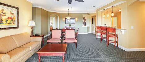A hotel room with a sofa, dining table, chairs, a ceiling fan, and a painting on the wall.