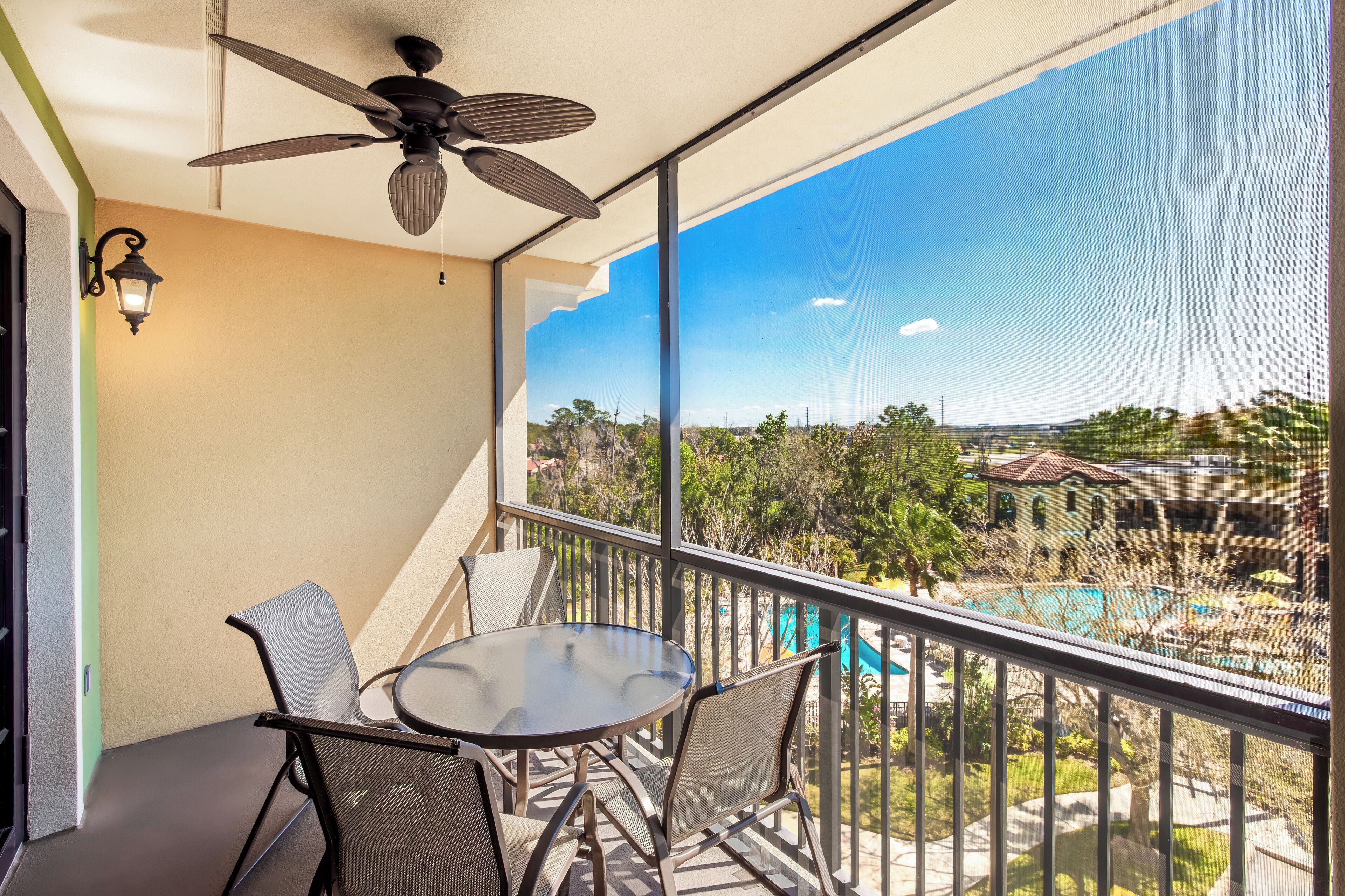A balcony with a table and chairs, a ceiling fan, and a view of a swimming pool and buildings.