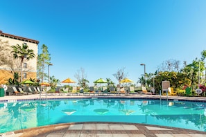 A swimming pool with lounge chairs and umbrellas, surrounded by palm trees and a building.