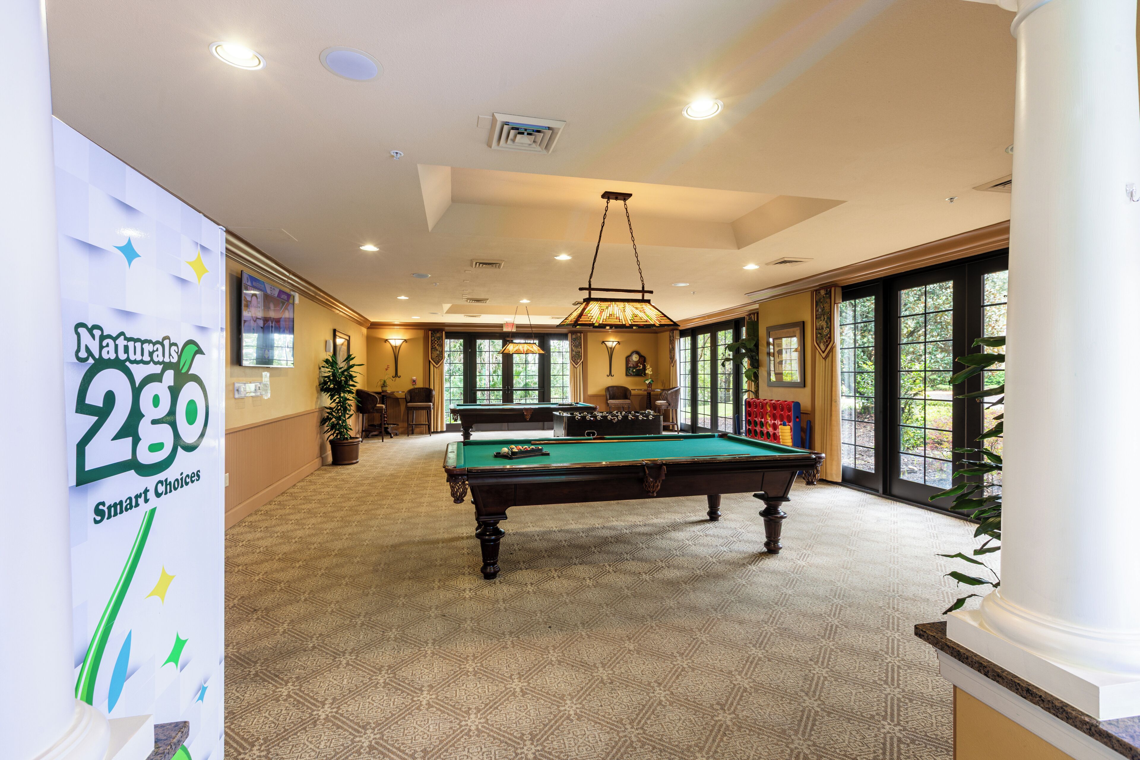 A pool table in a well-lit room with a patterned carpet, a large window, and a sign for Naturals 2GO Smart Choices.