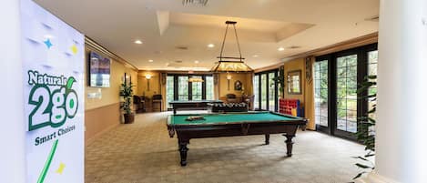 A pool table in a well-lit room with a patterned carpet, a large window, and a sign for Naturals 2GO Smart Choices.