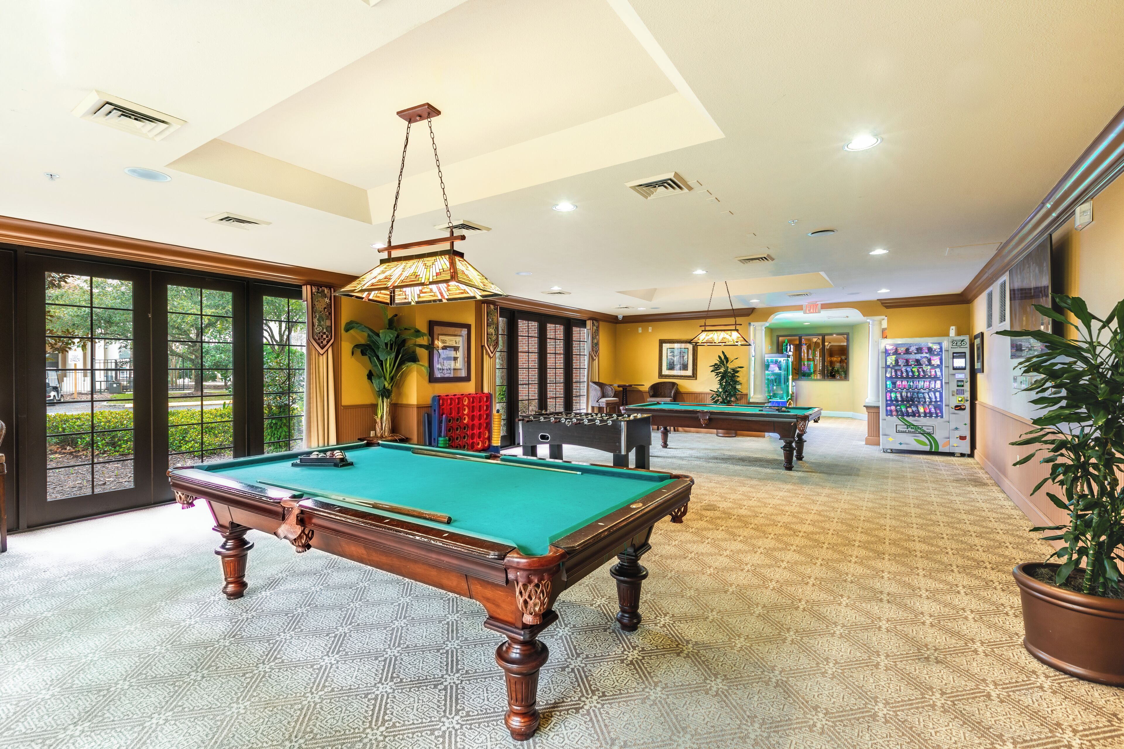 A pool table in a well-lit room with a patterned carpet, a billiards table in the background, and a vending machine.