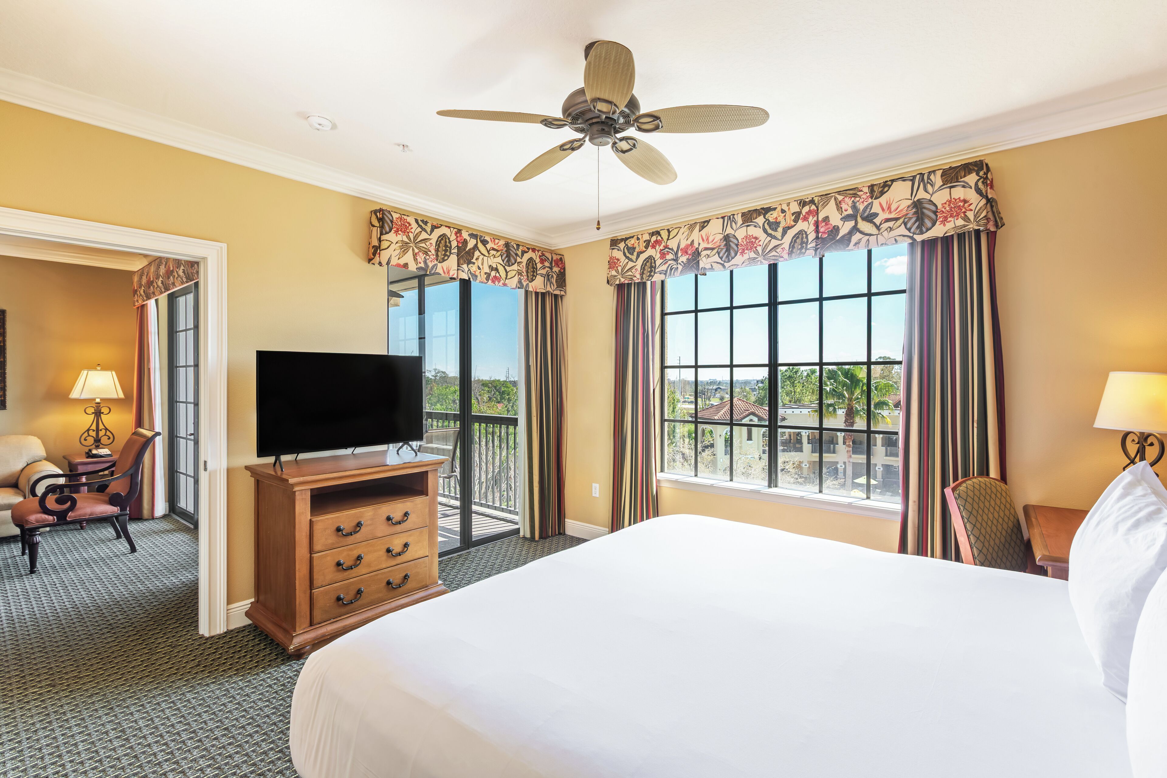 Een hotelkamer met een groot bed, een flatscreen televisie, een houten dressoir, een balkon met uitzicht en een plafondventilator.