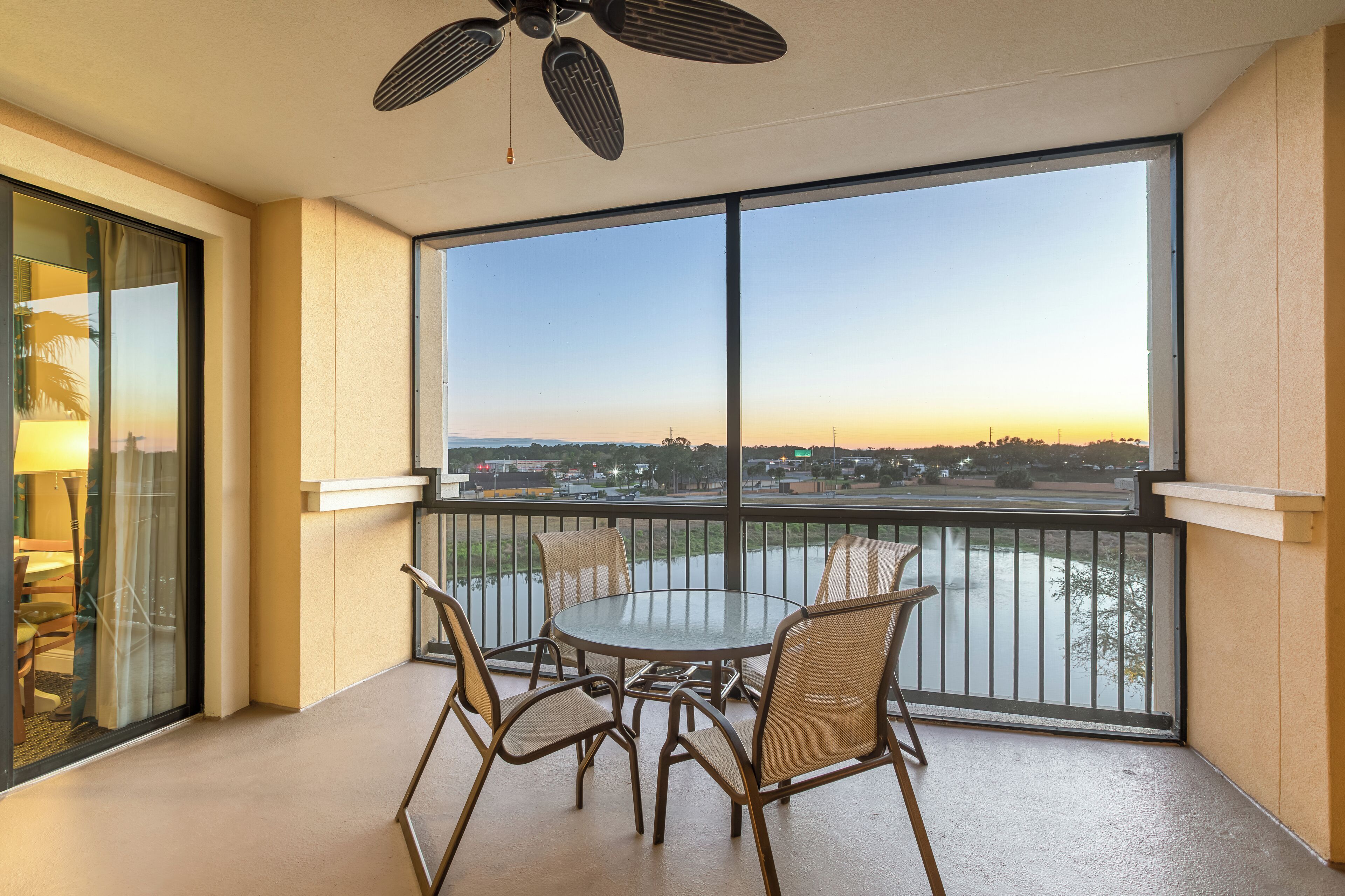 Un balcon avec une table et des chaises en verre, un ventilateur de plafond et une vue sur une étendue d’eau et le paysage urbain au loin.
