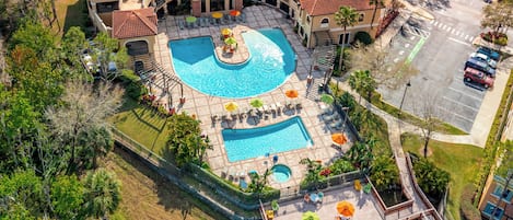 An aerial view of a resort with a large swimming pool, multiple lounge areas with umbrellas, and a parking lot.
