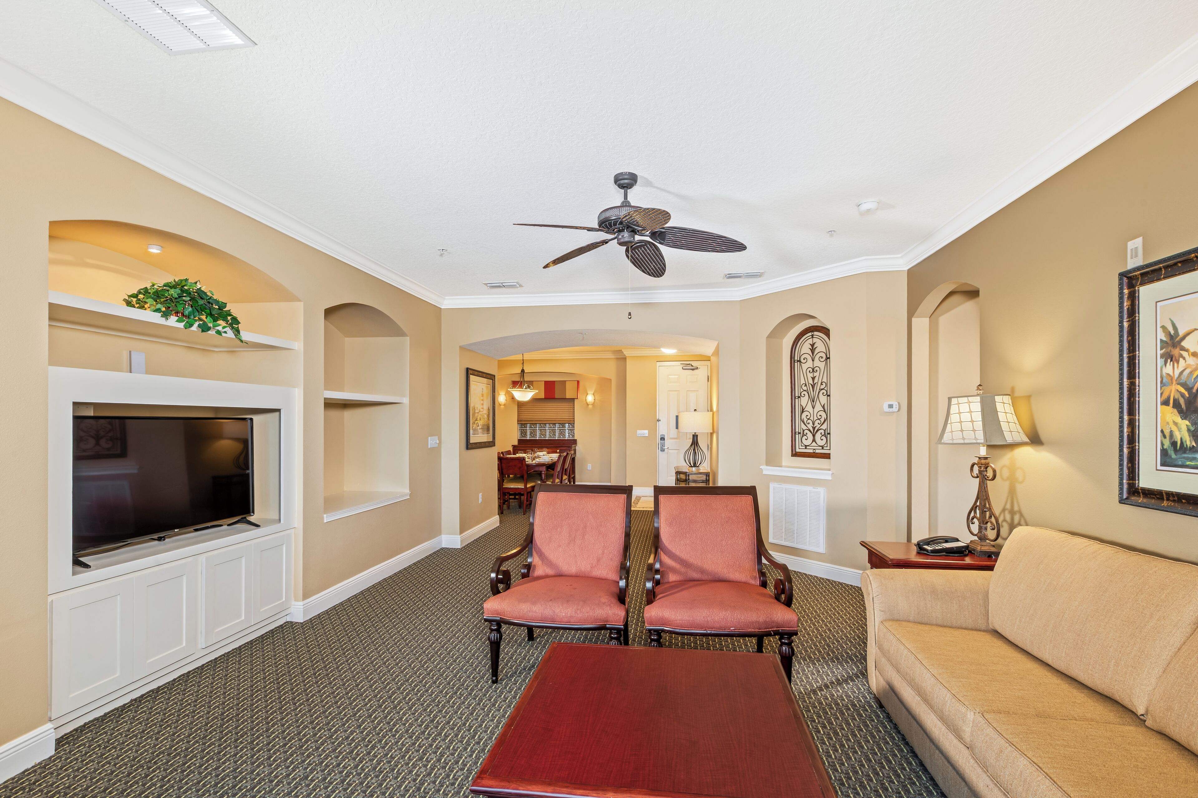 A hotel living room with a flat-screen TV, two armchairs, a sofa, a coffee table, a ceiling fan, and a dining area in the background.