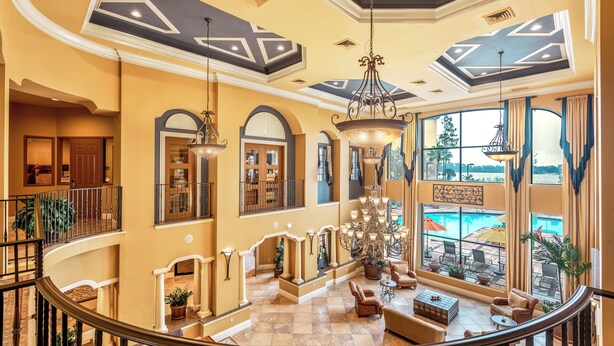 A spacious lobby with a grand chandelier, large windows offering a view of a pool, and a second-floor balcony with a railing.