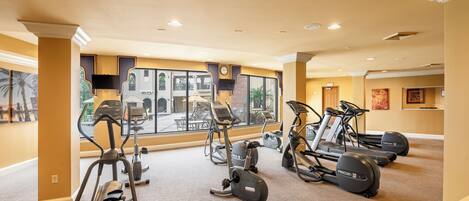A gym with treadmills, elliptical machines, and stationary bikes, large windows, and a view of the outside.