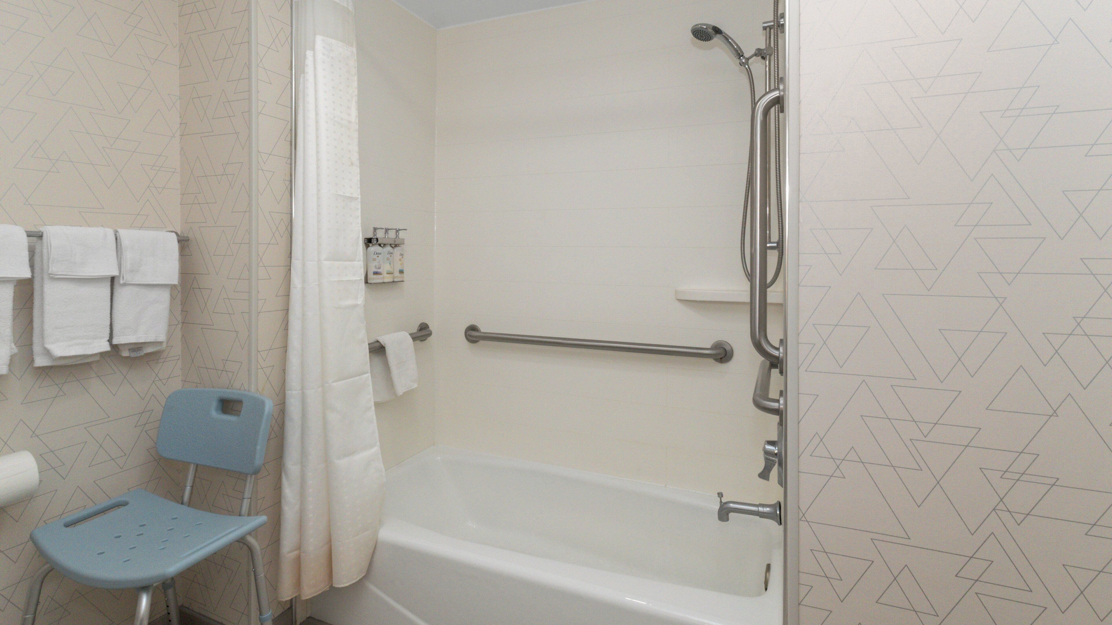 A bathroom with a bathtub, a shower, a handrail, a towel rack, and a blue chair.