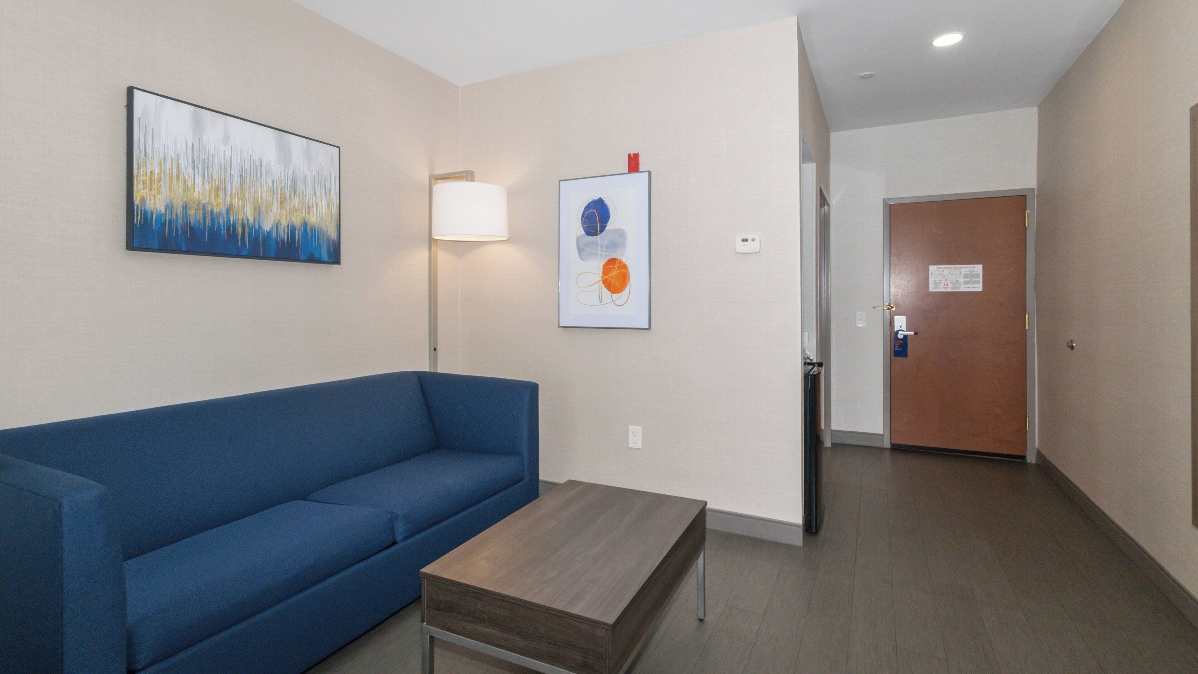 A hotel room with a blue sofa, a wooden coffee table, and two wall-mounted artworks.