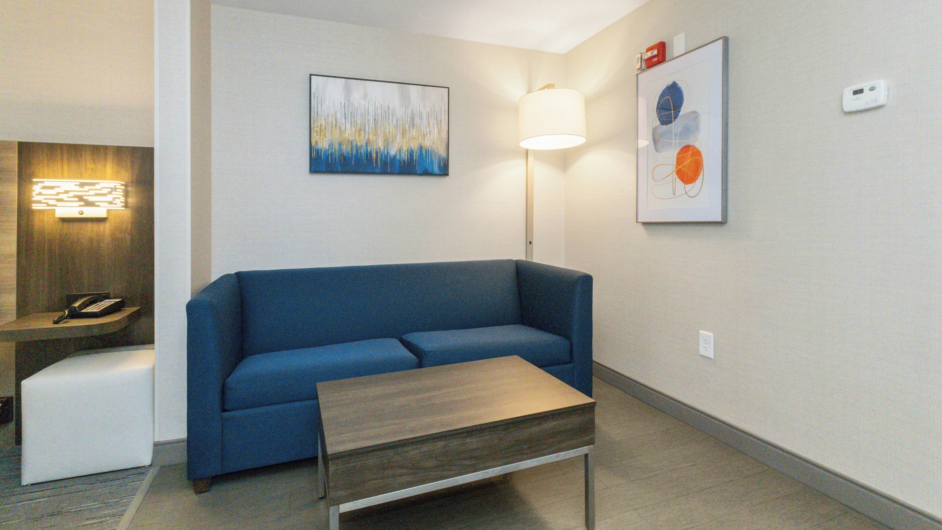 A modern hotel room with a blue sofa, a wooden coffee table, a desk with a lamp, and two abstract paintings on the walls.