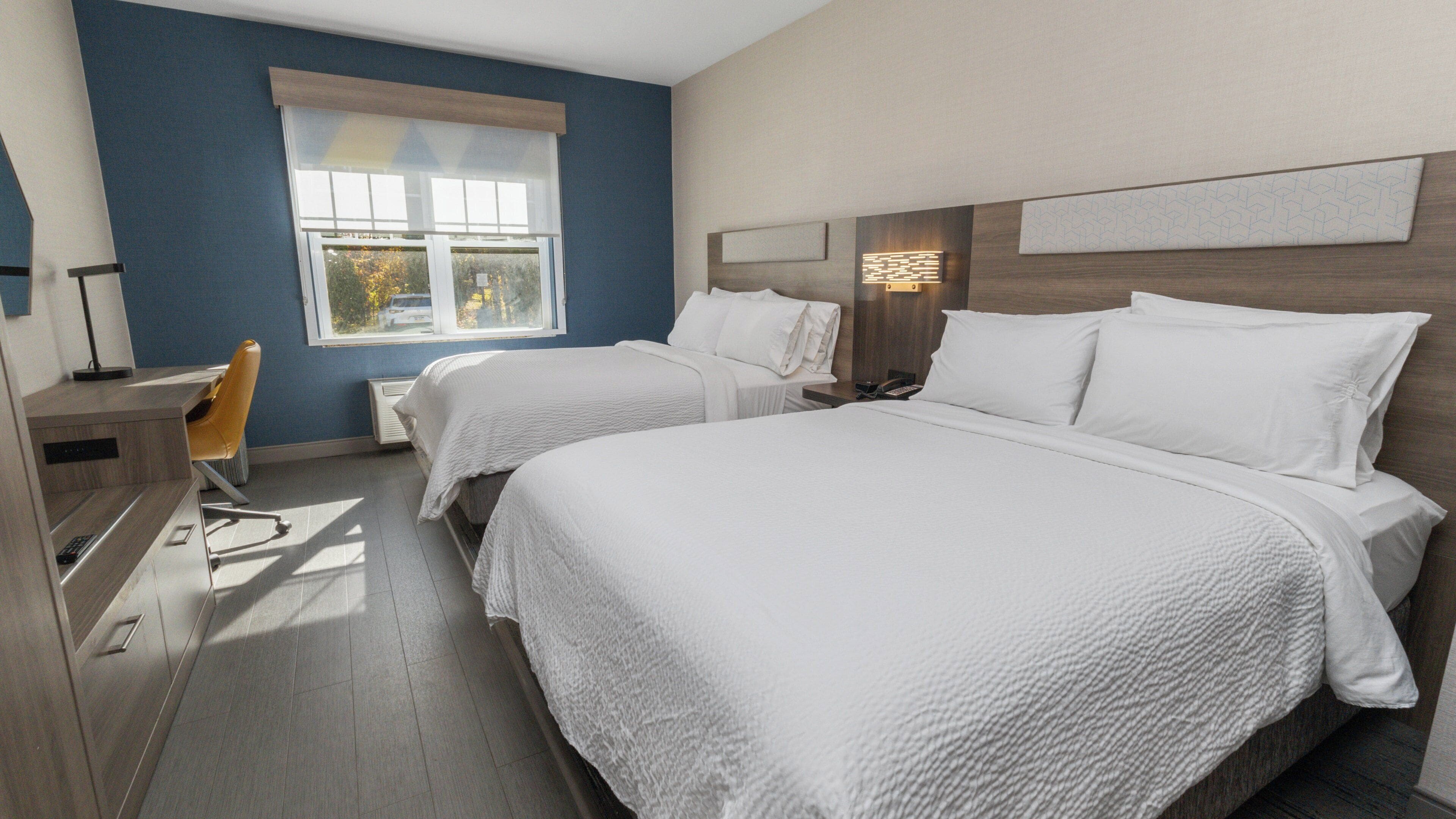 A hotel room with two beds, a desk, a chair, and a window.
