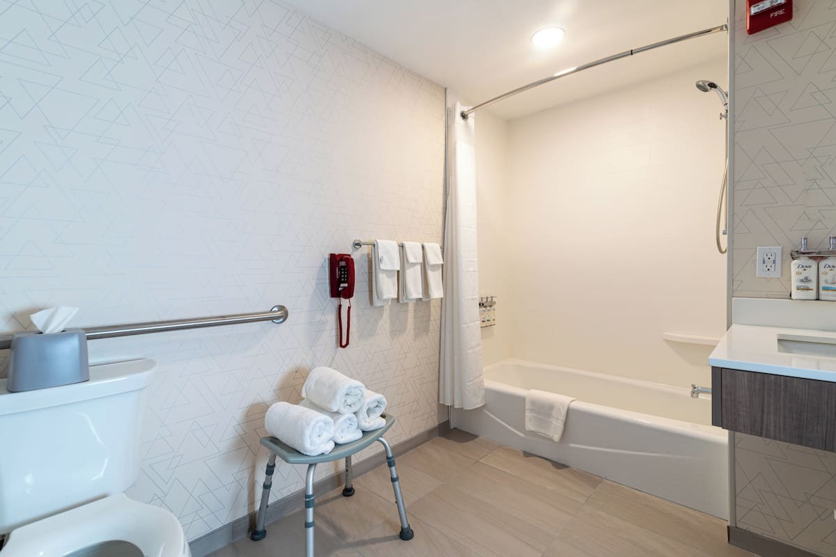 A modern bathroom with a toilet, a shower area, and a bathtub. There is a handrail, a towel rack with towels, and a small table with folded towels.