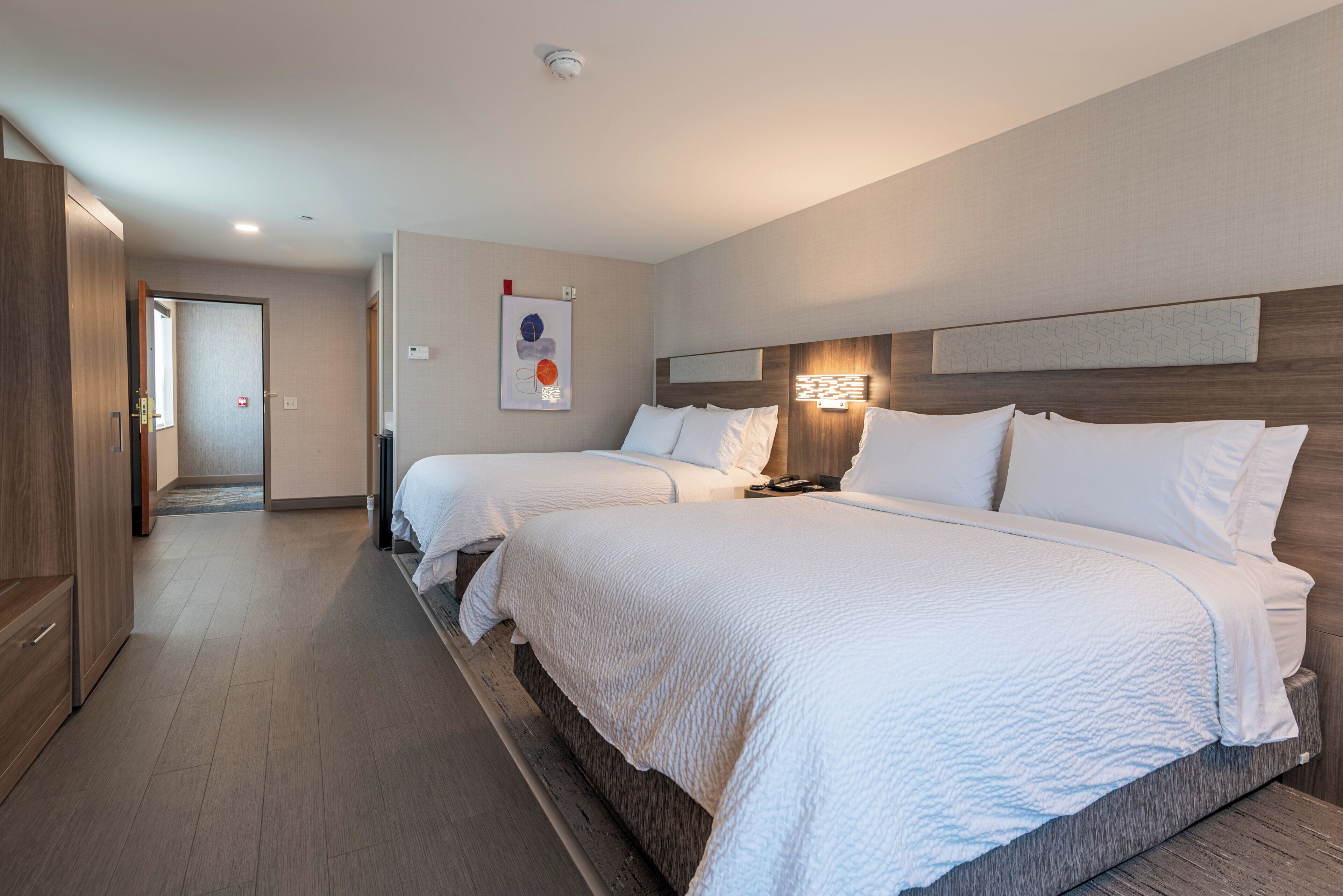 Habitación de hotel con dos camas, cabecera de madera, una obra de arte en la pared y una mesita de noche con lámpara.