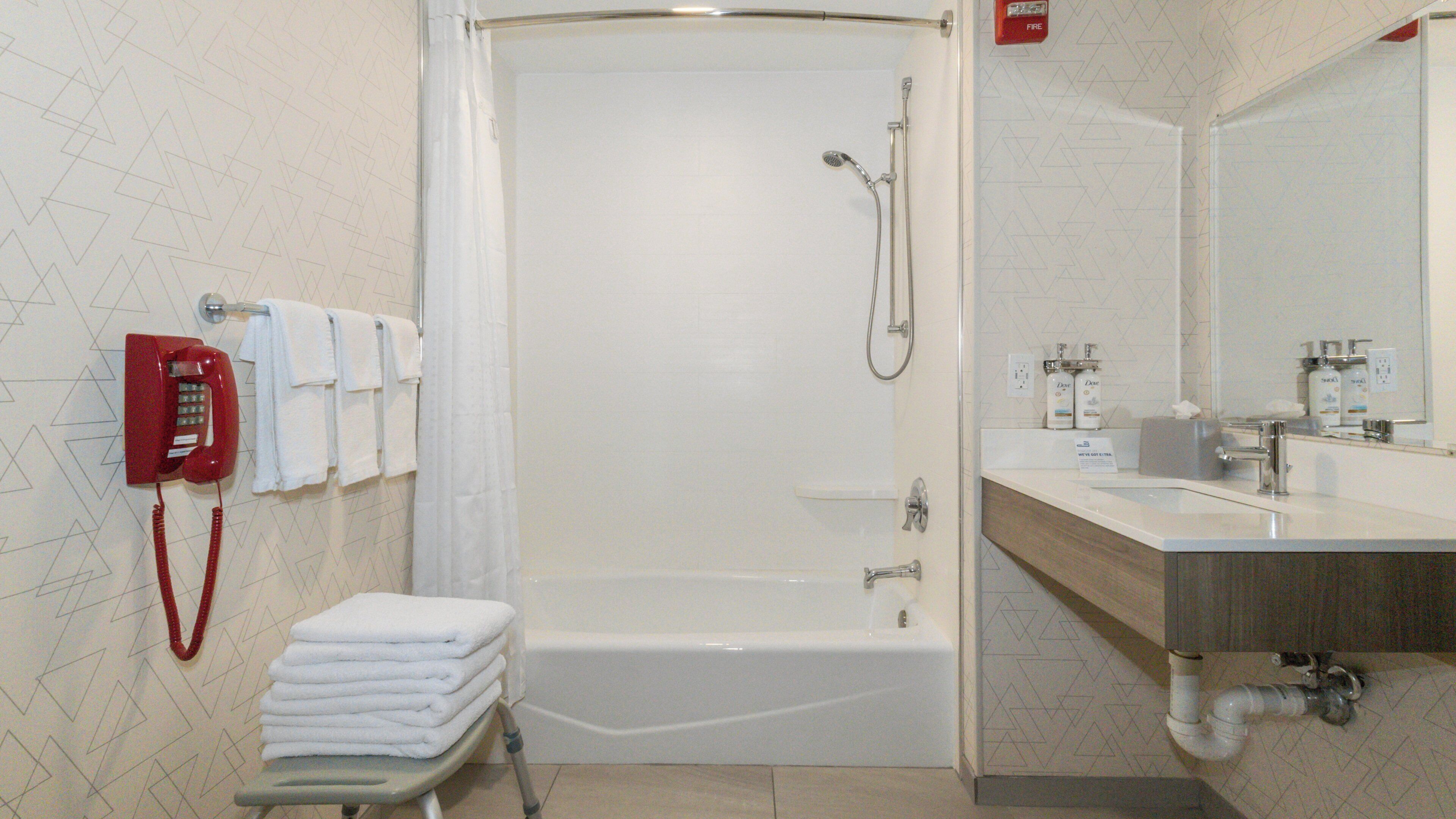 A modern bathroom with a shower, toilet, and sink. There is a red telephone on the wall and a stack of white towels.