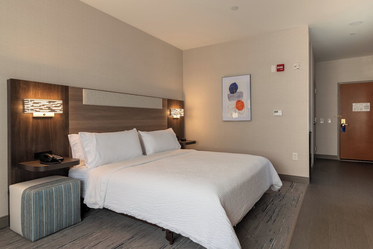 A modern hotel room with a large bed, a bedside table with a phone, a wall-mounted light, and a small bench.