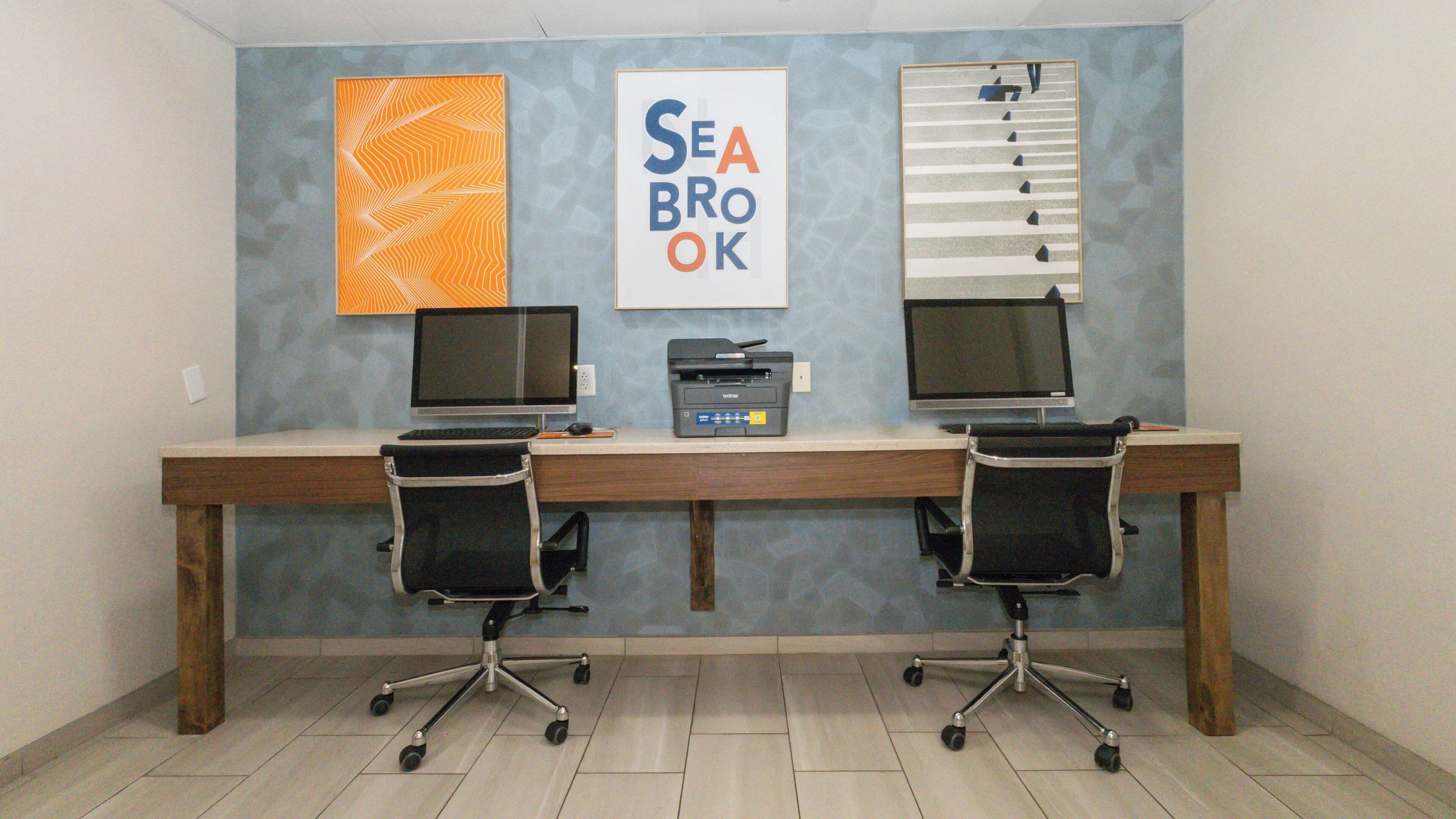 A workspace with two desks, each with a computer monitor, keyboard, and printer. There are two office chairs, a wall-mounted shelf, and two framed artworks.