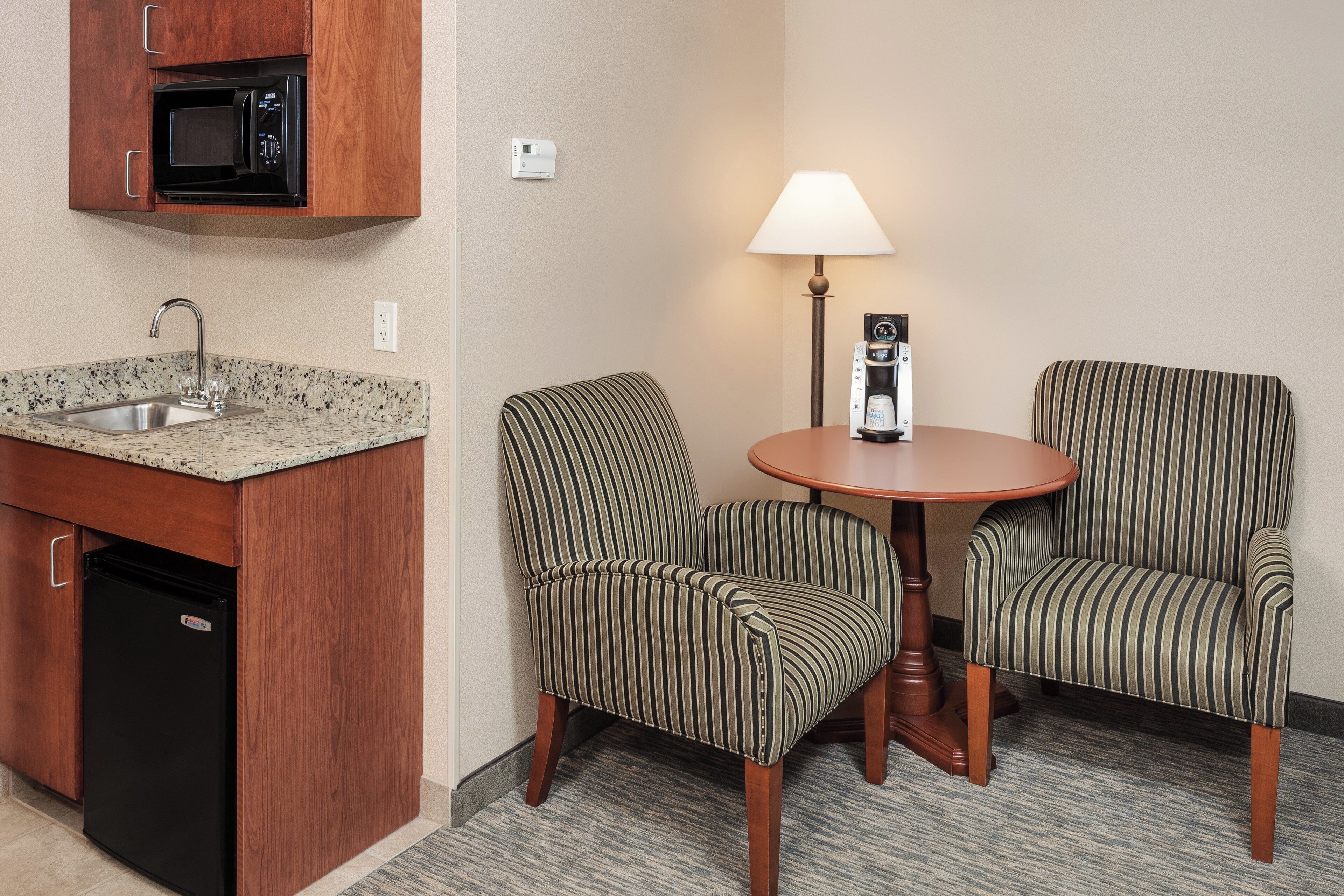 Una habitación de hotel compacta con kitchenette, dos sillones a rayas, una pequeña mesa redonda, una lámpara y una cafetera.