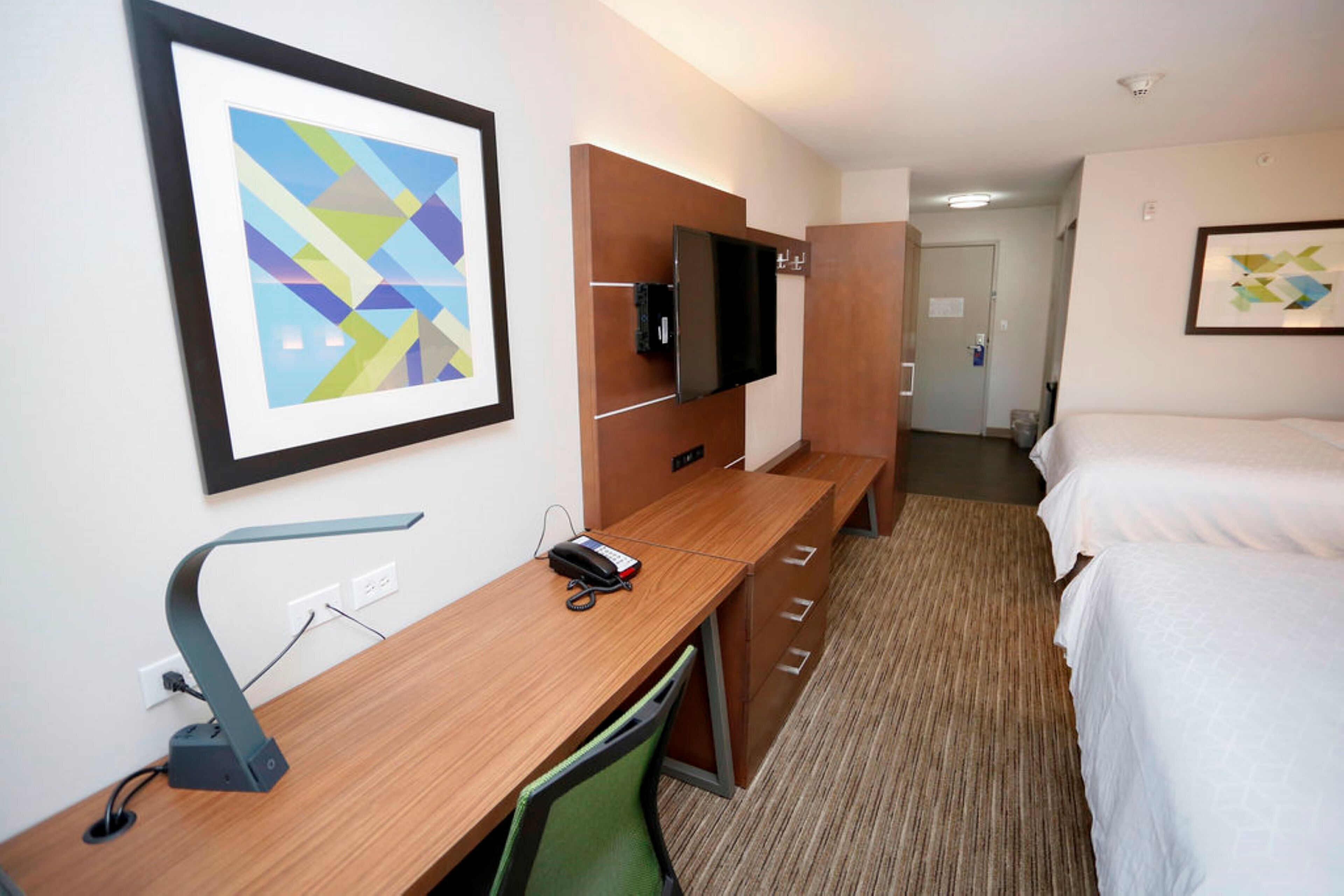 A hotel room with a wooden desk, a flat-screen TV mounted on a wooden panel, a bed with white bedding, and a framed abstract artwork on the wall.