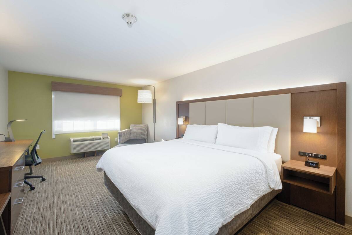 A hotel room with a large bed, a desk with a chair, a lamp, and a nightstand with a clock.