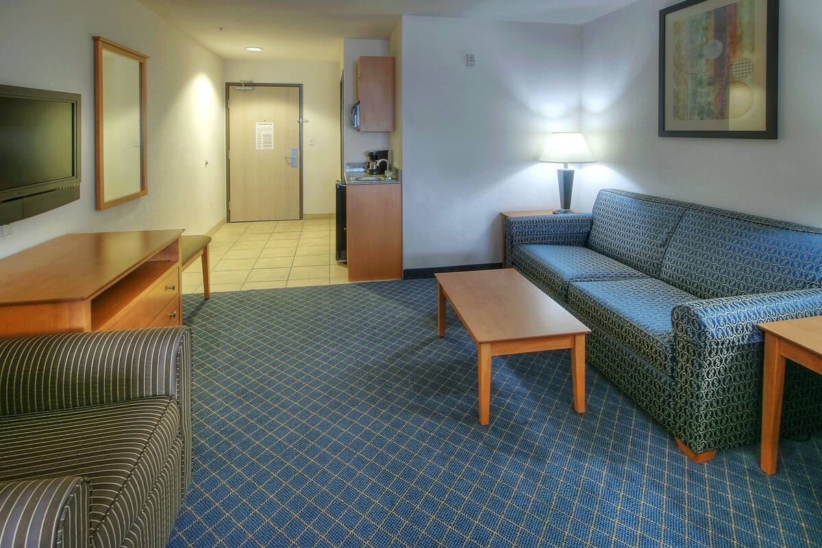 A hotel room with a sofa, a small table, a TV, a kitchenette, and a patterned carpet.