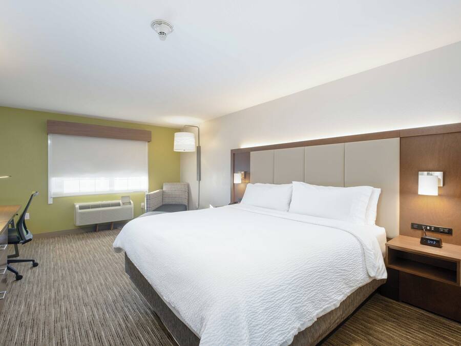 A hotel room with a large bed, a desk with a chair, a lamp, and a nightstand with a clock.