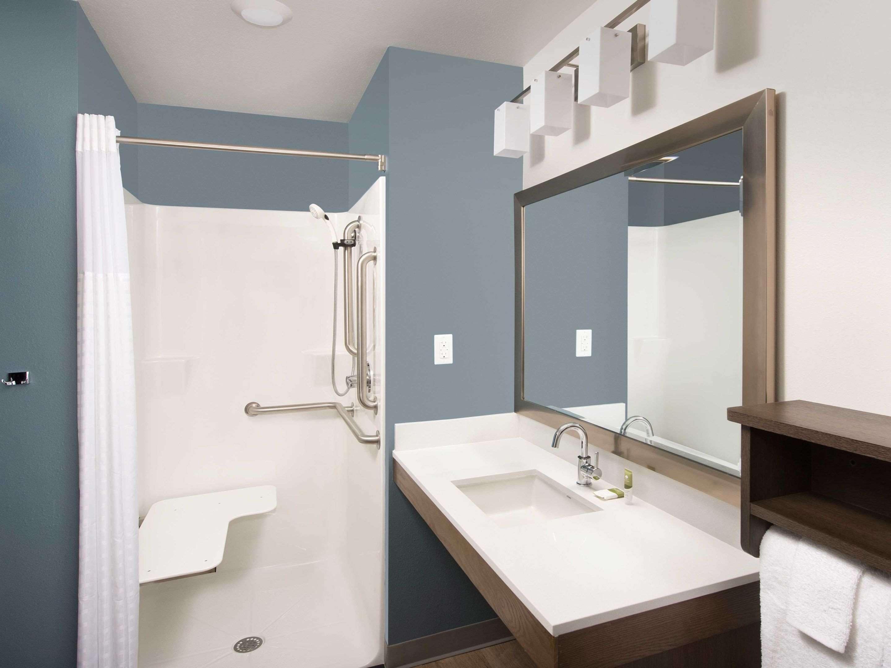 A modern bathroom with a walk-in shower, grab bars, a large mirror, and a vanity with a sink and towel rack.