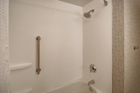 Un baño con paredes de azulejos blancos, una ducha con cabezal de ducha de mano, un barra de apoyo y una pared decorativa con mosaicos.