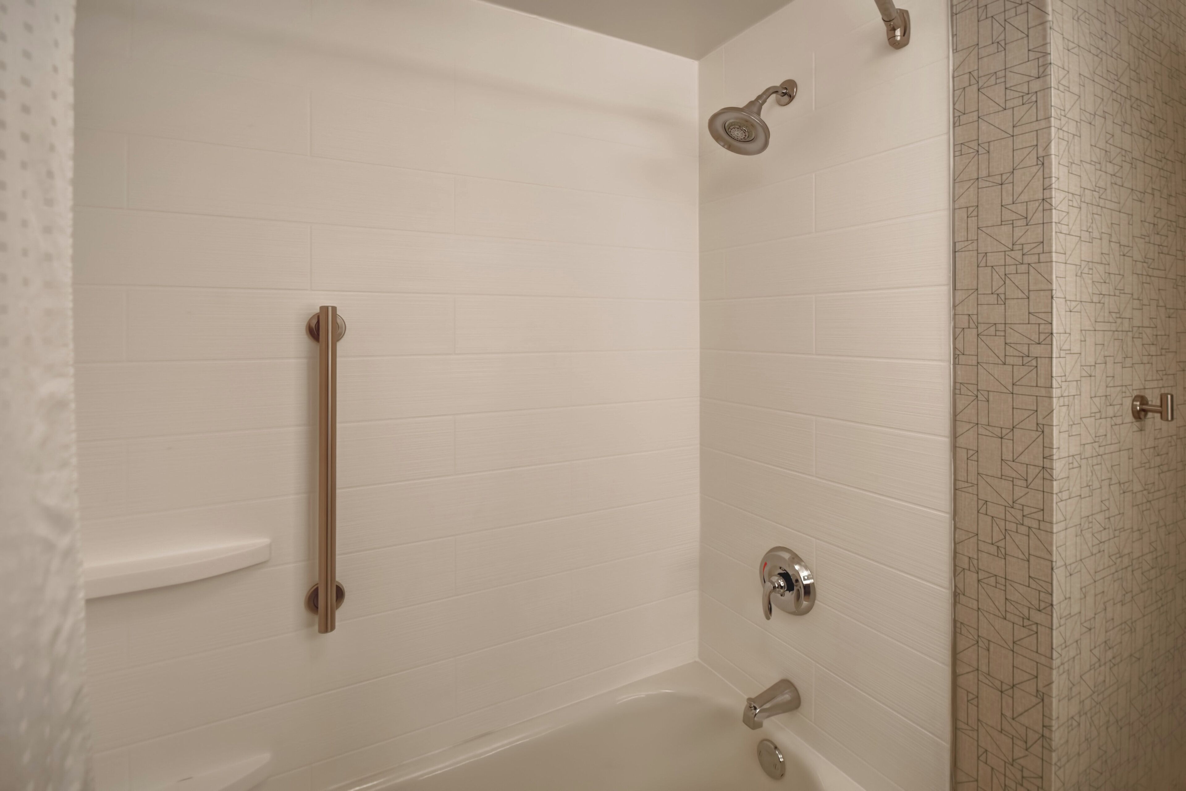 A bathroom with white tiled walls, a shower with a handheld showerhead, a grab bar, and a patterned tile accent wall.