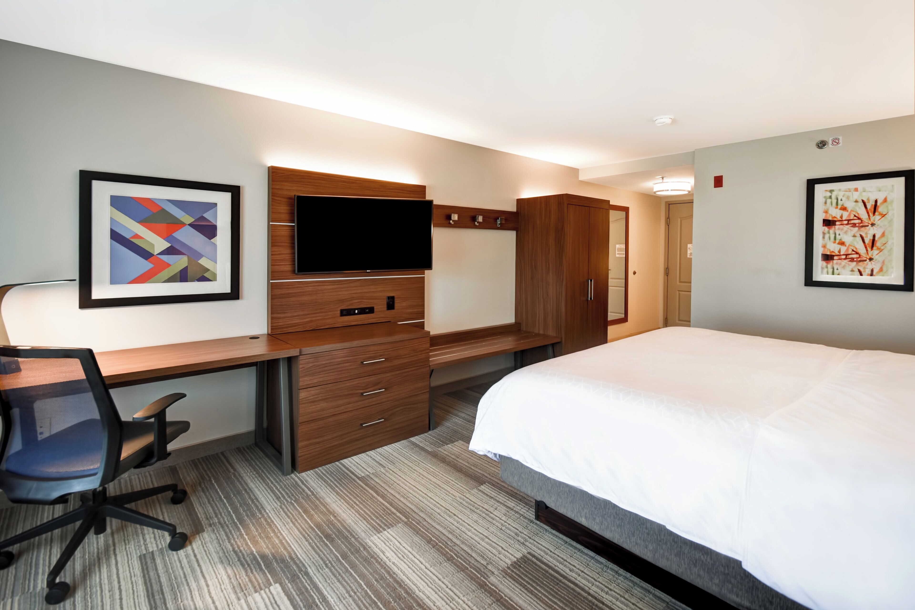 Una camera d'albergo con un letto, una scrivania, una sedia, televisione, armadio e opere d'arte.