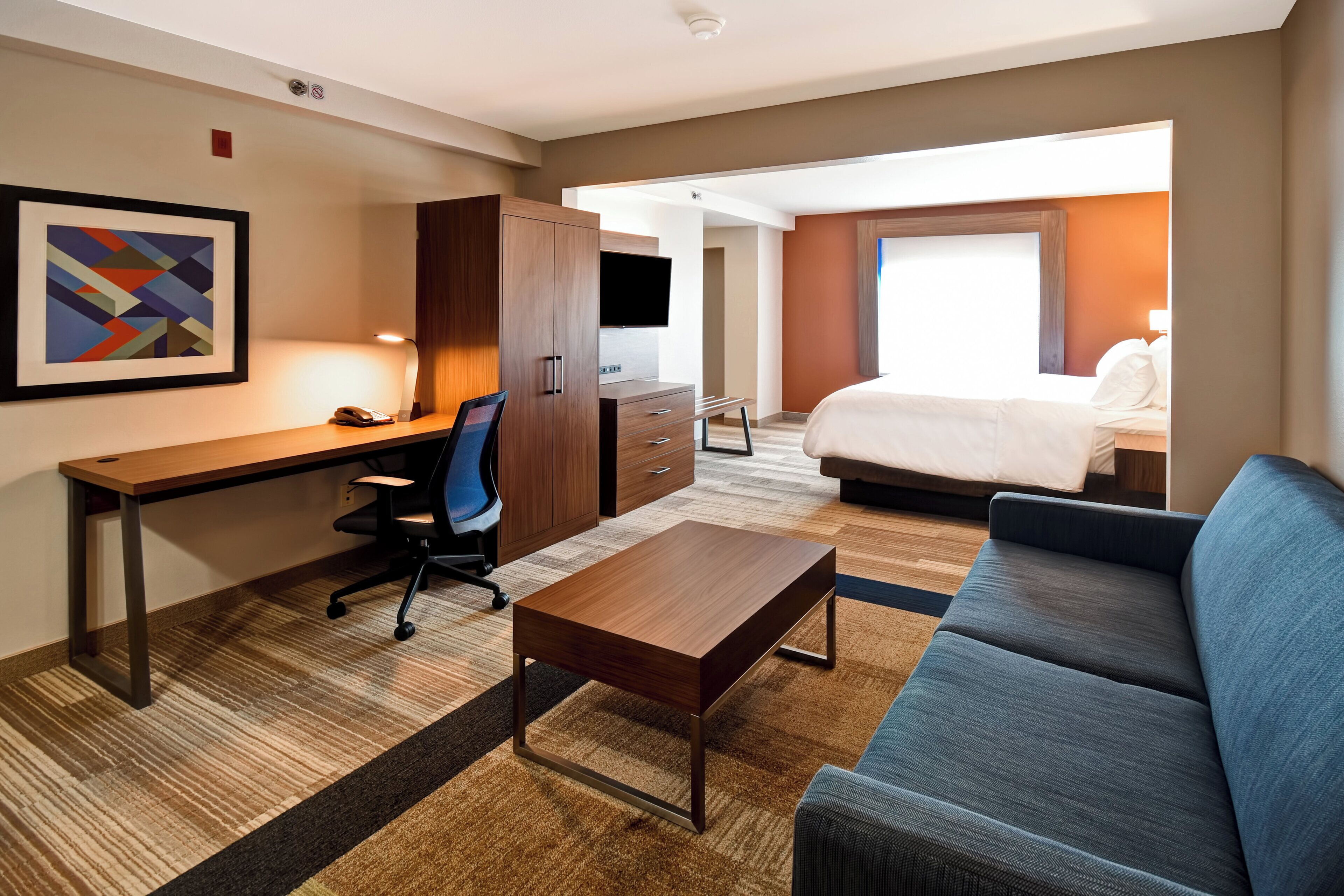 Une chambre d’hôtel avec un bureau, un canapé bleu, un lit, une télévision et un tableau encadré au mur.
