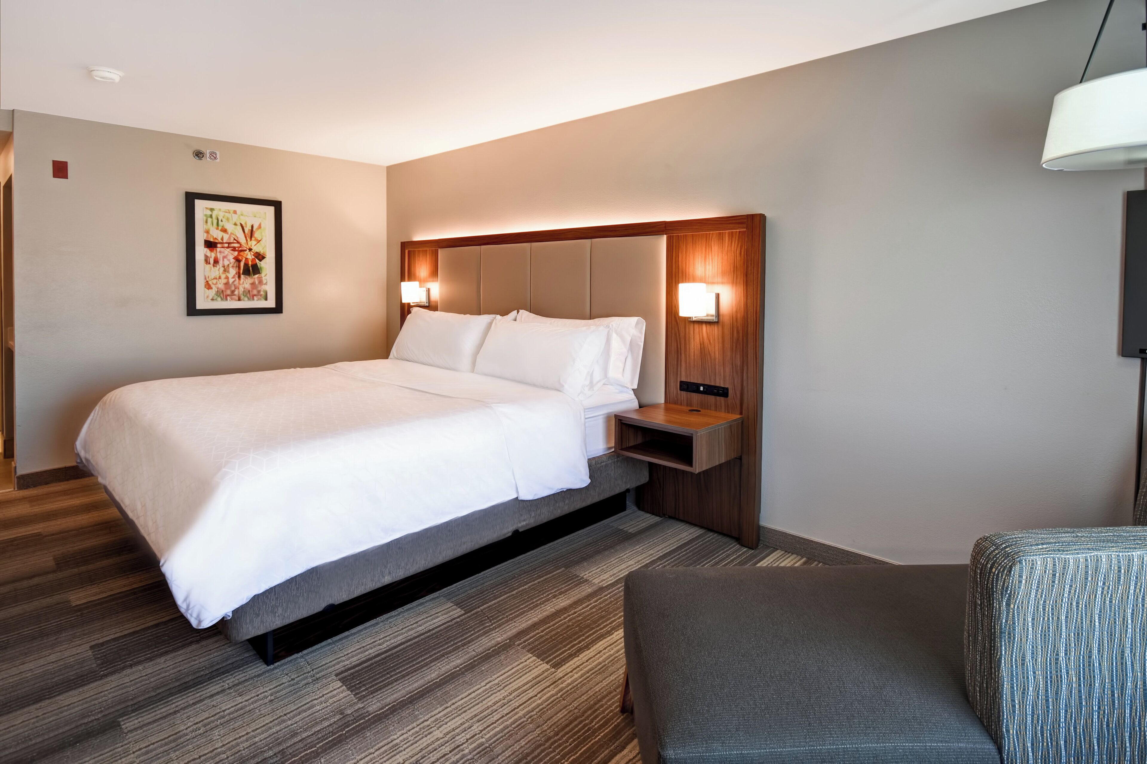 A hotel room with a bed, bedside table, lamp, and a framed picture on the wall.