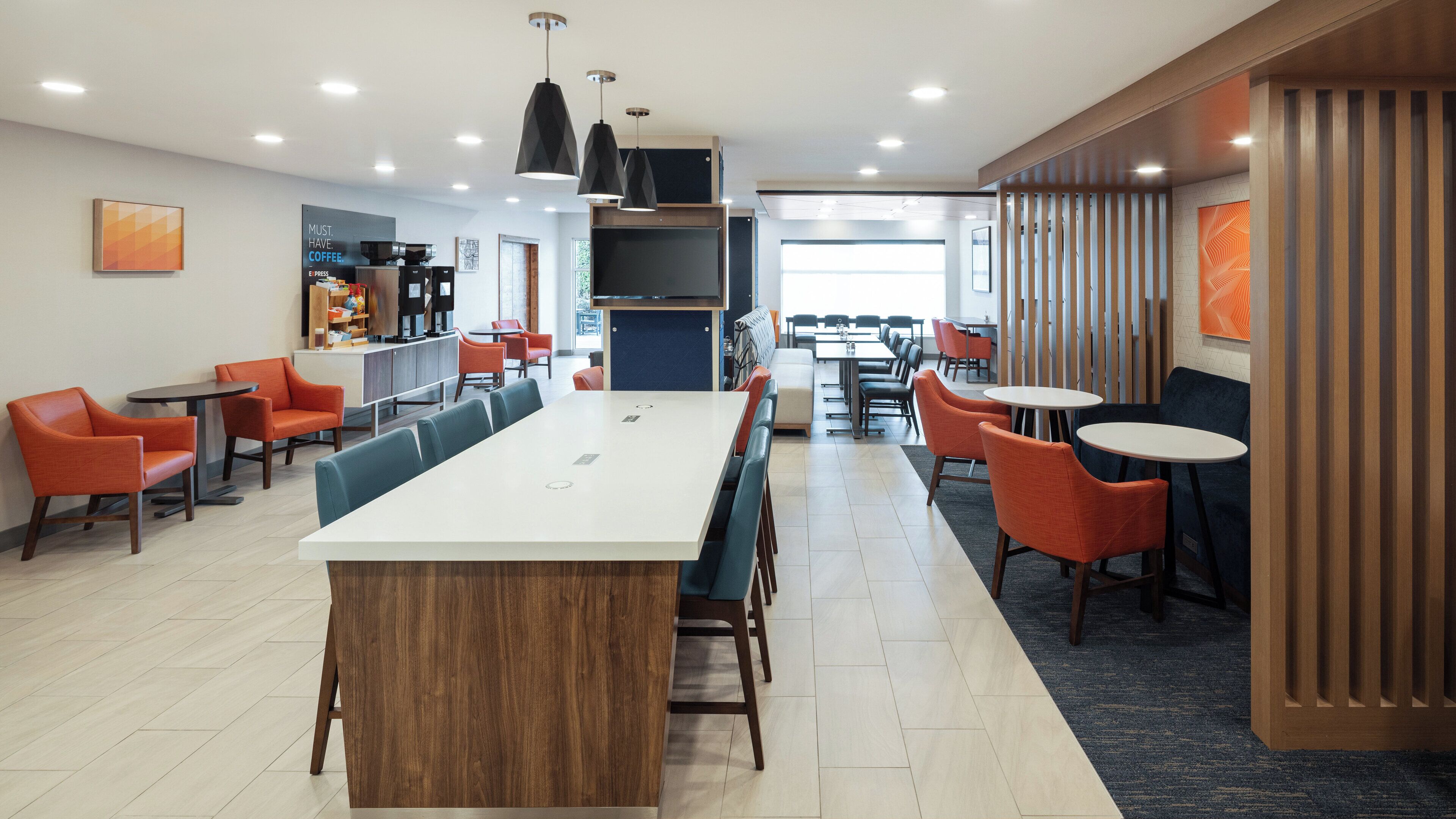 A modern hotel lobby with a long communal table, orange and blue chairs, a coffee station, and a view into the dining area.