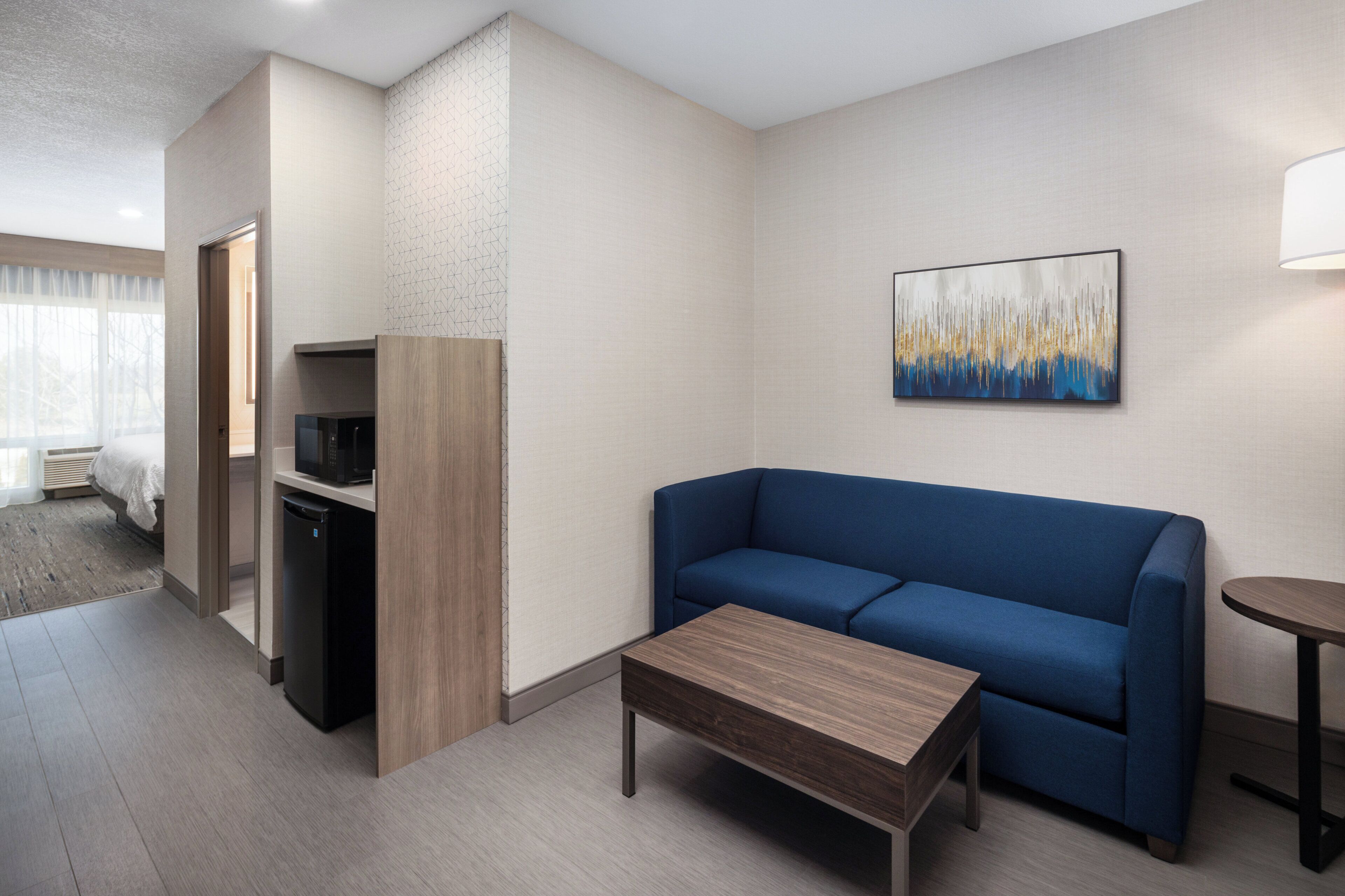 Una habitación de hotel moderna con un sofá azul, una mesa de centro de madera, un televisor de pantalla plana y un cuadro en la pared.