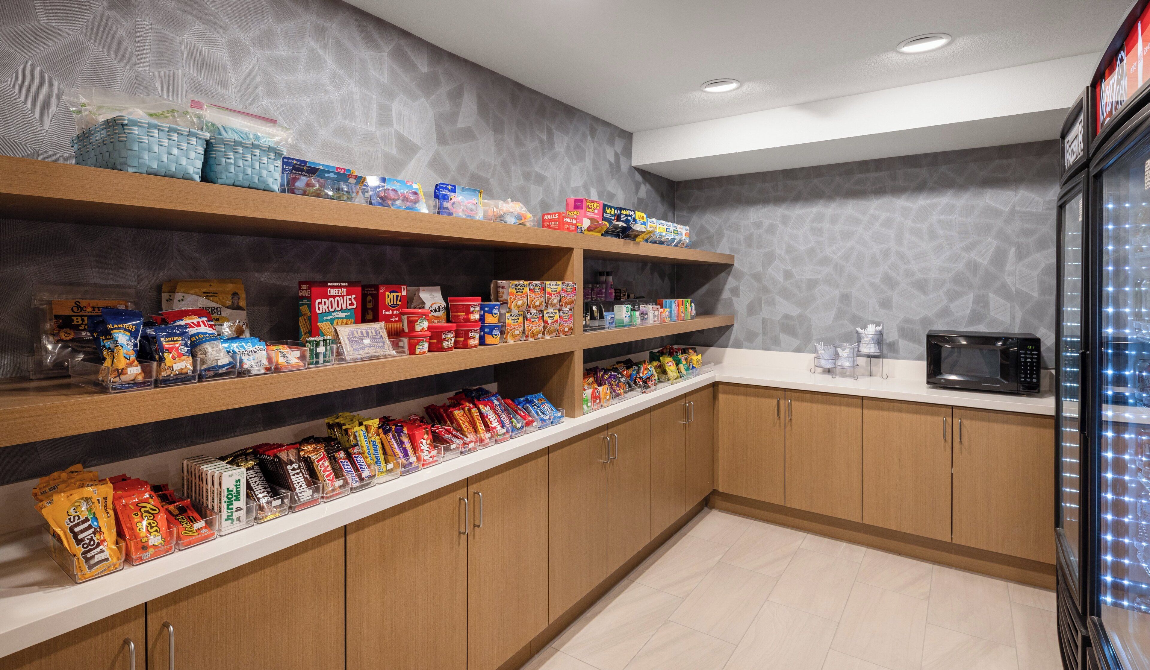 A kitchen area with wooden cabinets, a microwave, and shelves stocked with various snacks and food items.