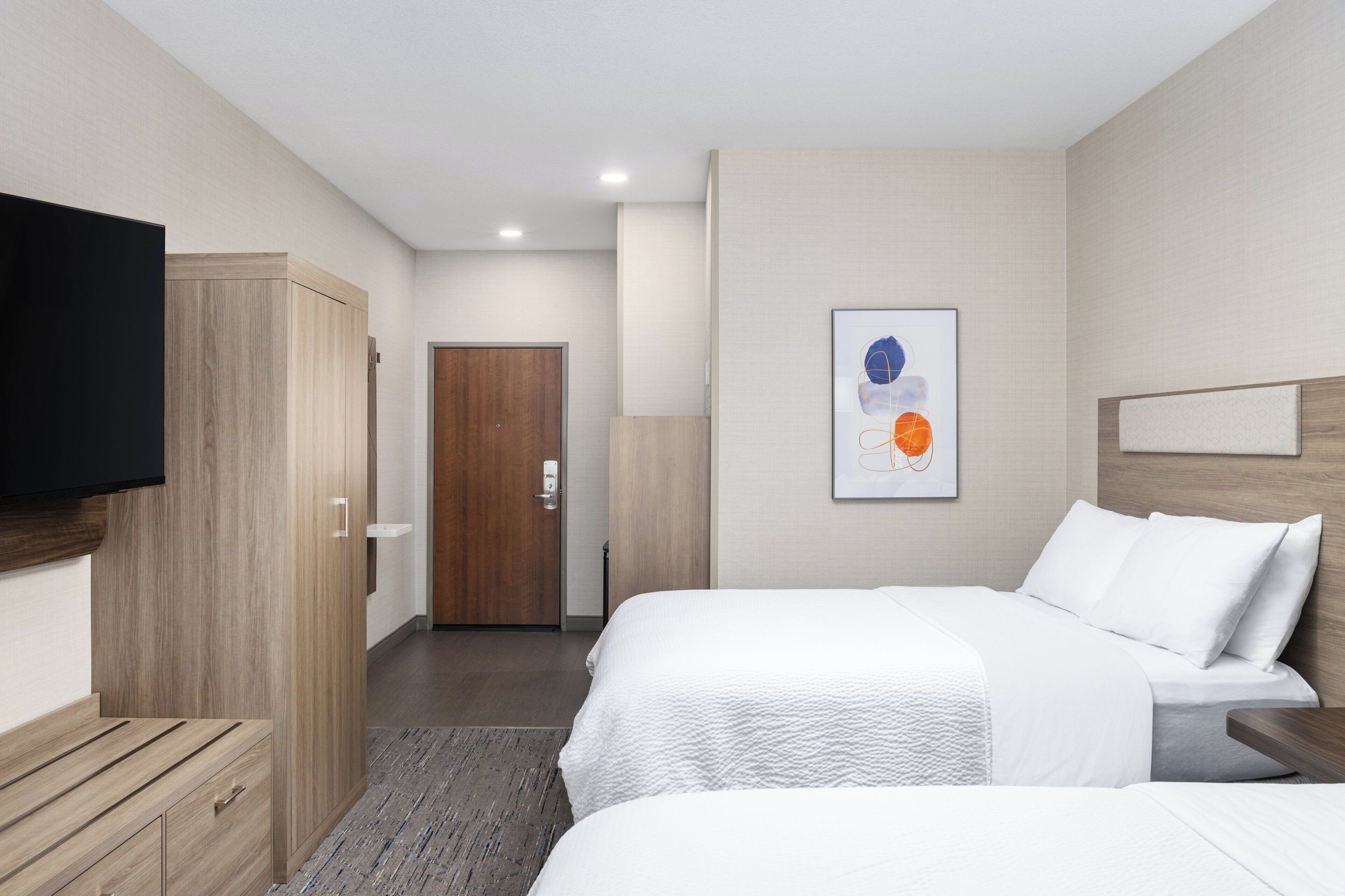 A hotel room with two beds, a TV mounted on the wall, a wooden wardrobe, and a door leading to another room.