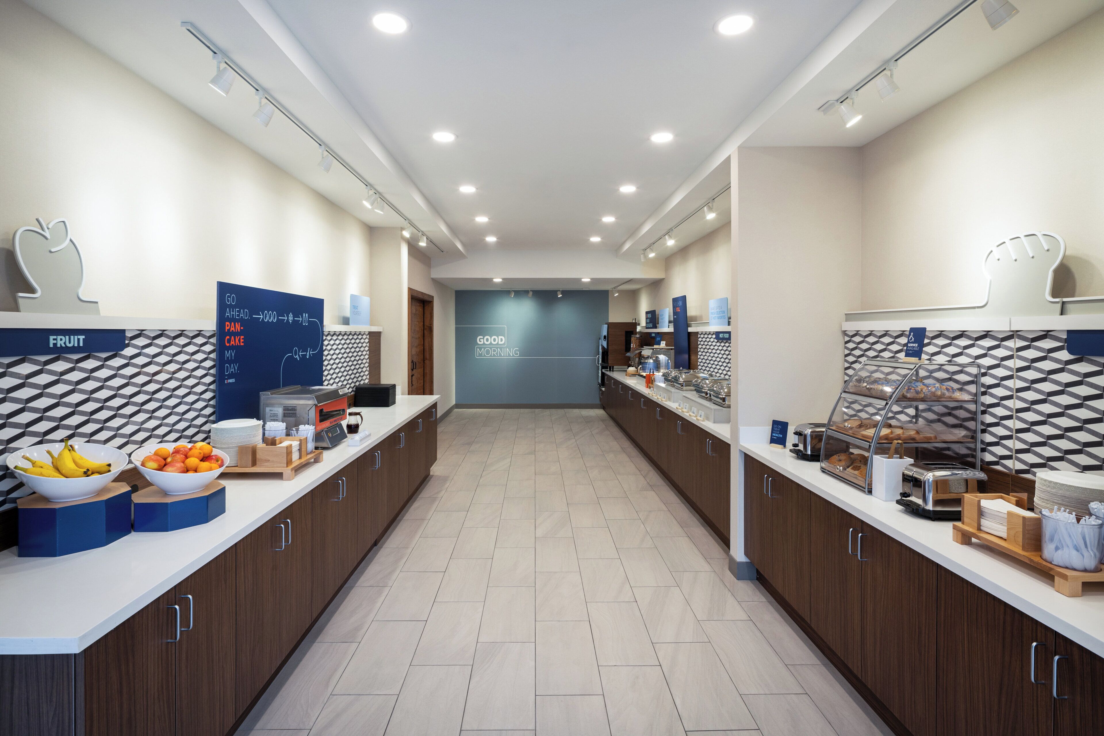 Un espace moderne dédié au petit-déjeuner, avec un long comptoir, des corbeilles de fruits et un panneau indiquant : « BONJOUR ».