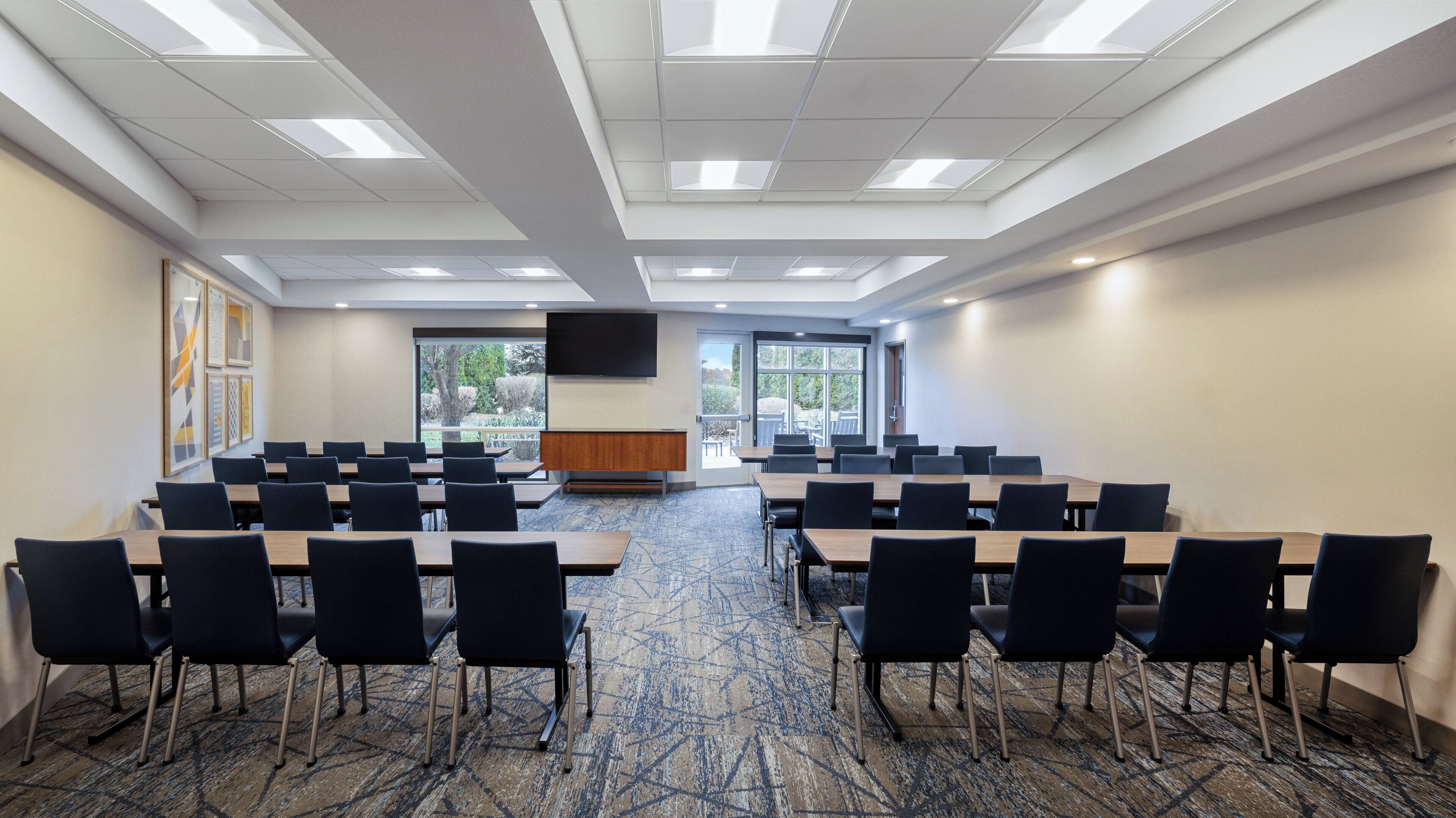 A conference room with multiple tables and chairs, large windows, and a flat-screen TV on the wall.