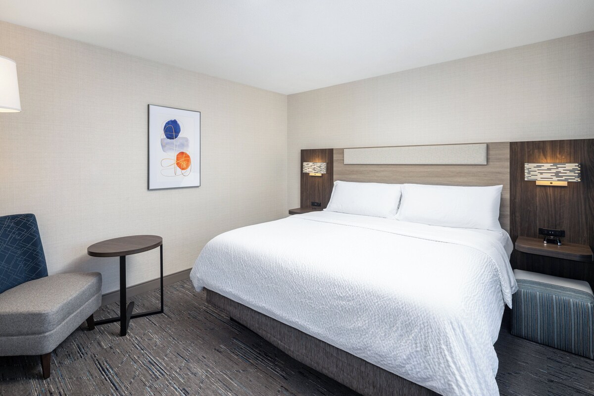 Quarto de hotel com uma cama grande, uma mesa pequena redonda, uma cadeira e uma obra de arte na parede.