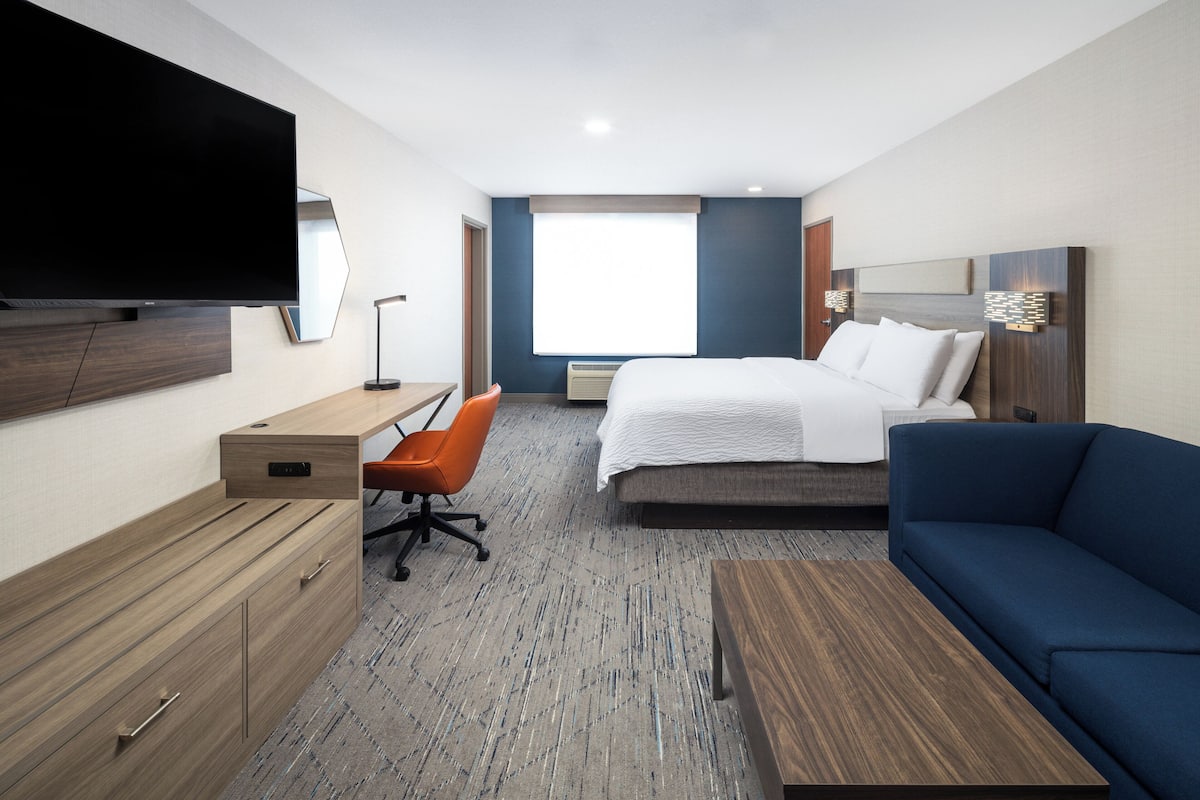 Una camera d'albergo con un letto, una scrivania con una sedia, una TV a parete e un'ampia finestra che lascia entrare la luce naturale.