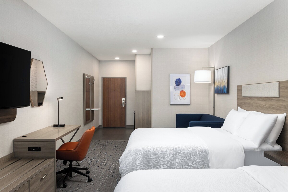 Quarto de hotel com duas camas, uma escrivaninha com cadeira, televisão na parede e uma porta que dá para outro cômodo.