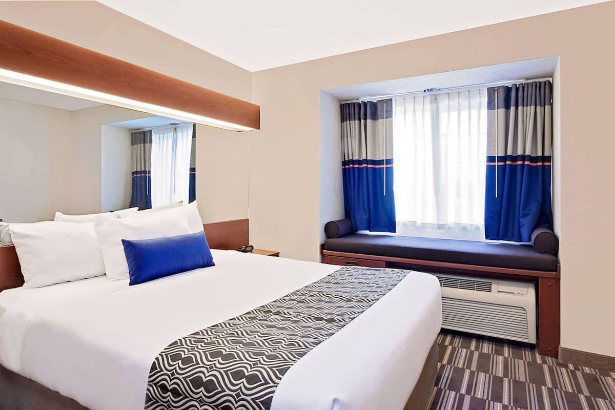 A hotel room with a large bed, a bedside table, a window with curtains, a built-in bench, and a wall-mounted air conditioning unit.