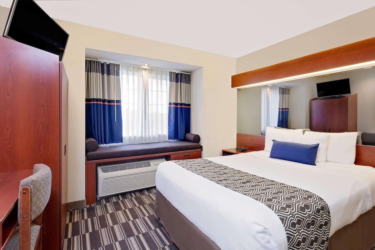 A hotel room with a bed, a desk, a chair, a window with curtains, a television, and a mirror.