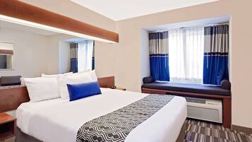 A hotel room with a large bed, a bedside table, a window with curtains, a built-in bench, and a mirror.