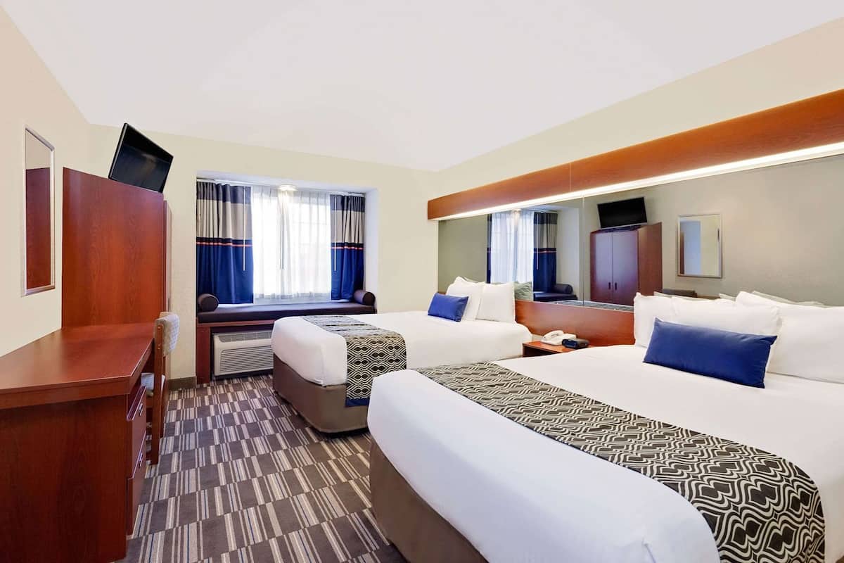 A hotel room with two beds, a desk, a chair, a mirror, a TV, and a window with curtains.