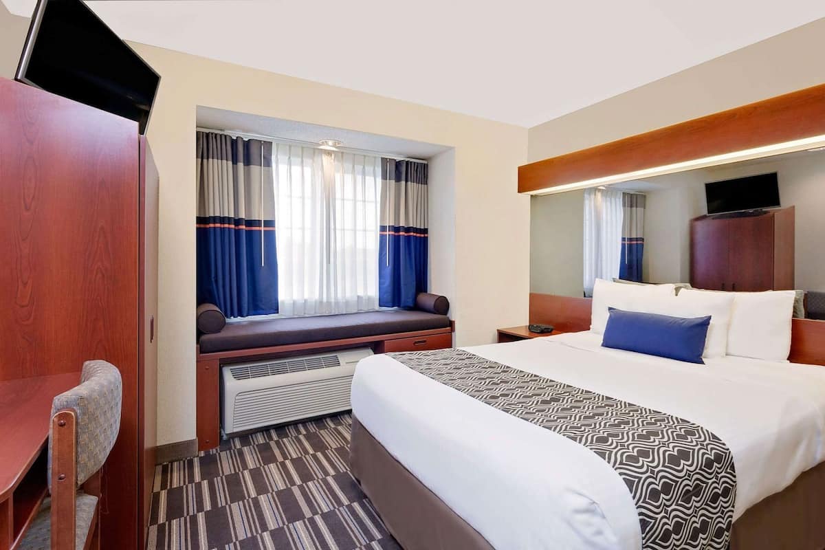 A hotel room with a bed, a desk, a chair, a window with curtains, a television, and a mirror.