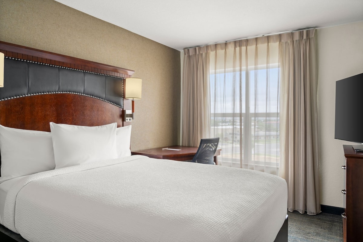 Habitación de hotel con una cama grande, un escritorio, una silla, un televisor y una ventana con cortinas.