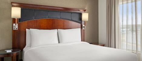 Una habitación de hotel con una cama grande, cabecera de madera, dos lámparas de noche y una ventana con cortinas translúcidas.